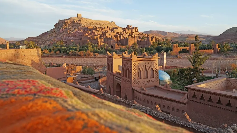 Sunset over an ancient fortified village with earthen buildings and a hilltop fortress, surrounded by greenery.